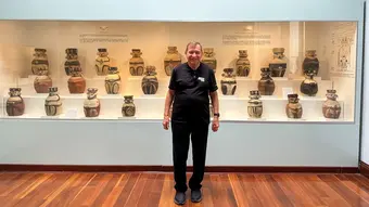 mężczyzna ubrany w ciemne ubrania stoi, za nim znajduje się gablota z ceramicznymi baniakami