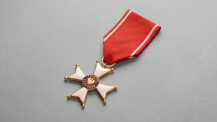 Złoty order w kształcie krzyża z rozszerzającymi sią na zewnątrz ramionami. Ramiona pomalowane na biało. W środku krzyża koło z godłem i napisem POLONIA RESTITUTA. Order wisi na czerwonej wstędze z dwoma białymi pasami po bokach. 