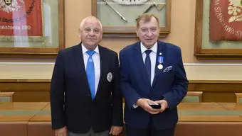 dwóch mężczyzn ubranych formalnie stoi obok siebie w sali i pozują do zdjęcia